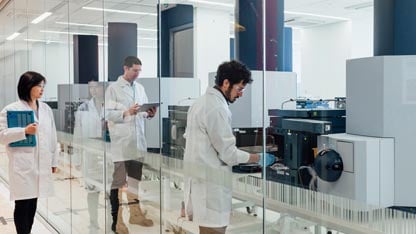 two scientists wearing white lab coats working in a laboratory facing a large rectangular analytical instrument 