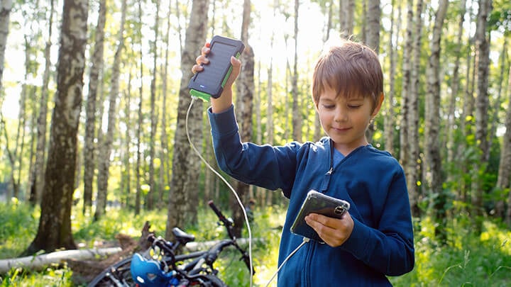 Young boy in a blue zipper jacket on a bike trail with a bike in the background holding up a small solar panel device connected to a phone