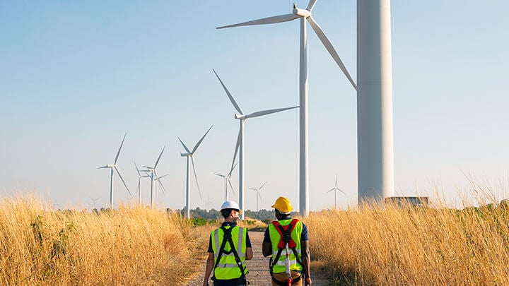 Two workers walking through a field with large wind turbines in the near distance