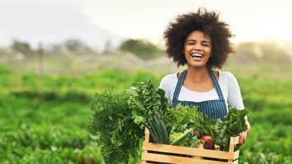 Smiling woman in an apron with wooden crate of fresh produce walking out of a field