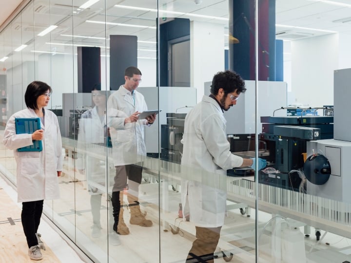 Two scientists can be seen working with large machinery through a clear glass wall in a laboratory