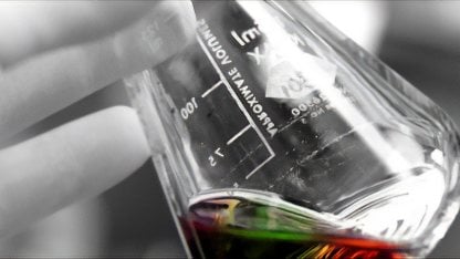 A hand coloured in shades of grey holding a glass chemical flask that contains a multi-coloured liquid