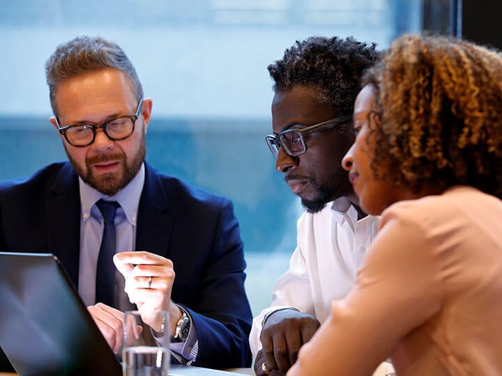 Business man in suit and tie wearing glasses talking to an African American couple looking at a laptop during a business meeting