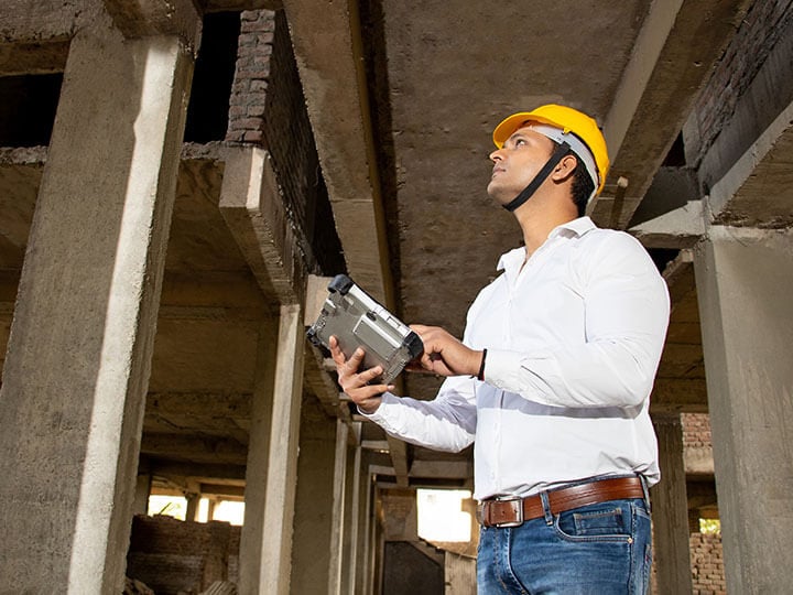 Young male architect wearing yellow hard hat, white button up shirt, and jeans, holding digital tablet inspecting a construction site structure