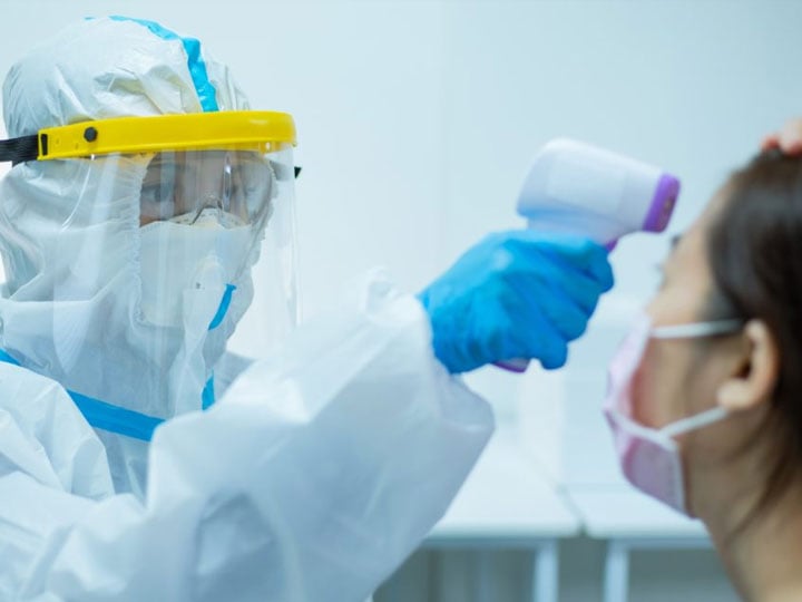 person in PPE equipment including full body suit, mask, and face shield checking temperature of a woman wearing a fabric facemask with a forehead thermometer