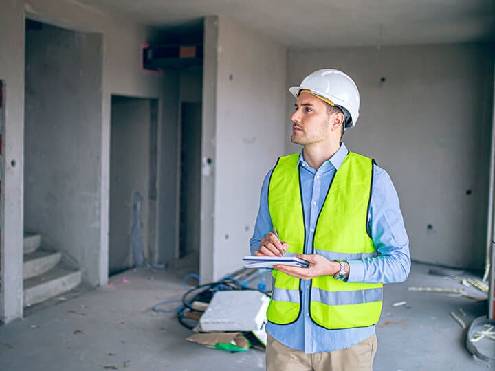 Mature male in white hard hat and yellow caution vest inspecting the interior of a construction site with a clipboard