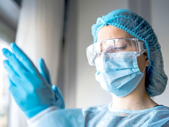 Medical professional wearing protective goggles, face mask and blue hair net, putting on protective blue gloves