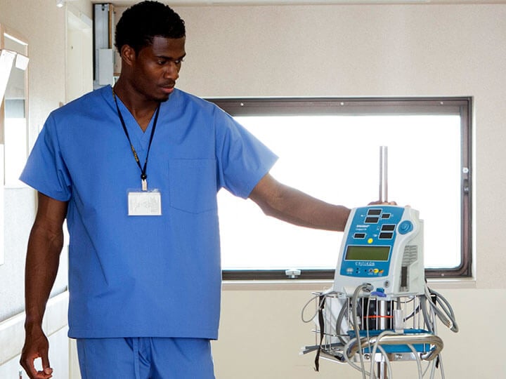 Male nurse in light blue scrubs walking down the hallway of a hospital looking at an medical device