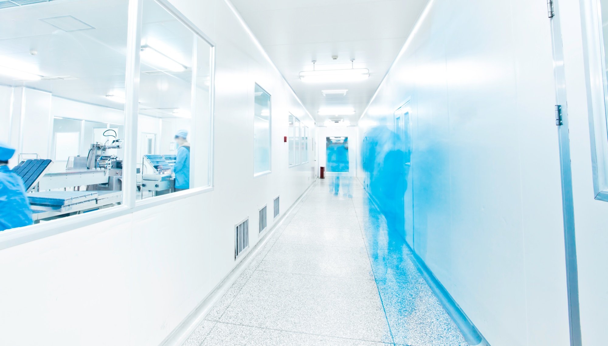 Workers in blue overalls walk down a sterile bright corridor in a pharmaceutical factory