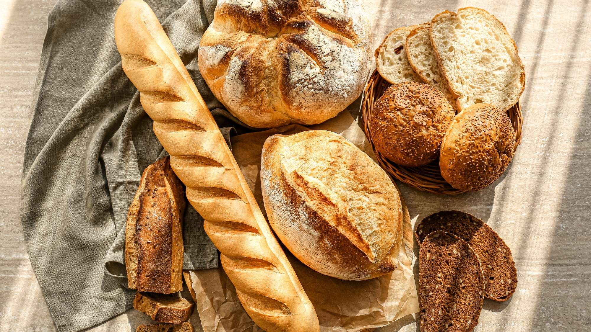 Close-up of a variety of bread, including a baguette, round sourdough, and seeded rolls, arranged on a linen cloth.